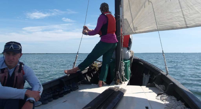 A group of students wearing life jackets sit and stand on a sailboat on calm blue water